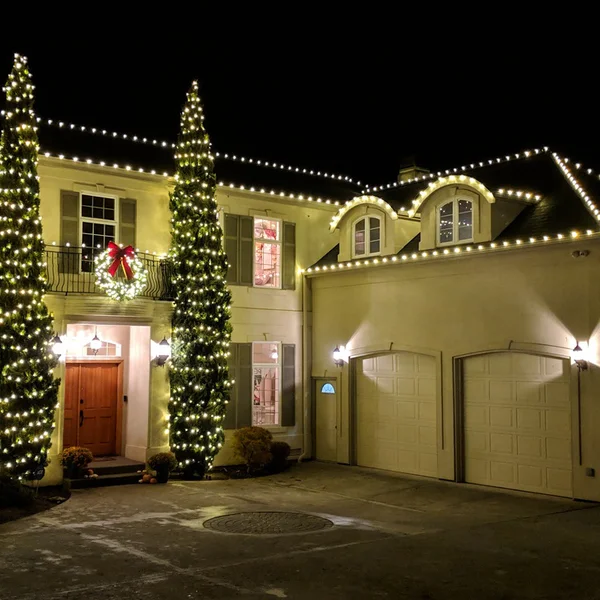 A beige two-story house is decorated with warm white Christmas lights along the roofline, trees, and a wreath on the balcony.