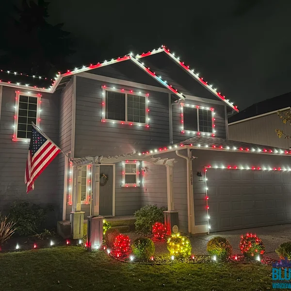 Gray two-story home adorned with red and white Christmas lights on the roofline, windows, and front yard bushes.