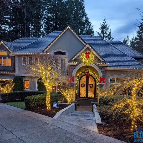 Residential home with professional Christmas light installation on roofline, trees, front door wreaths and festive bows.