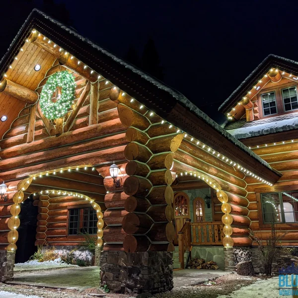 Festive log home illuminated with installed Christmas lights and a wreath near the roofline.