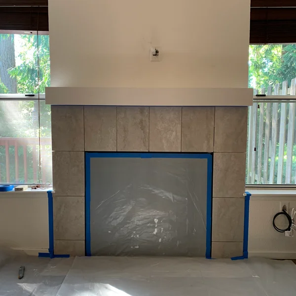 During a home remodel, a tiled fireplace in gray is partially covered with plastic and blue tape.