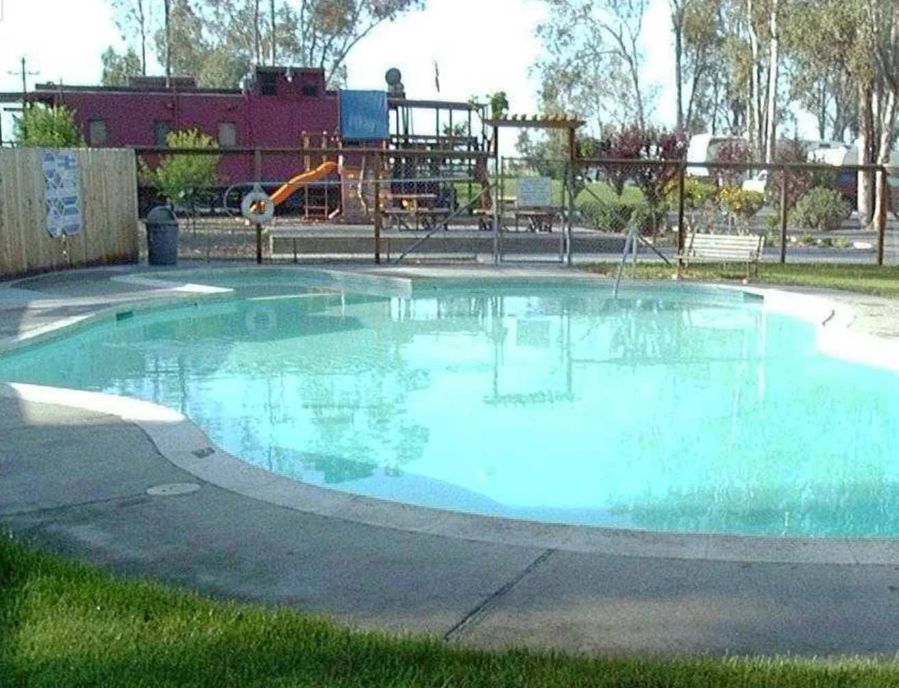Outdoor swimming pool at a manufactured home community, with a playground and red building visible in the background.