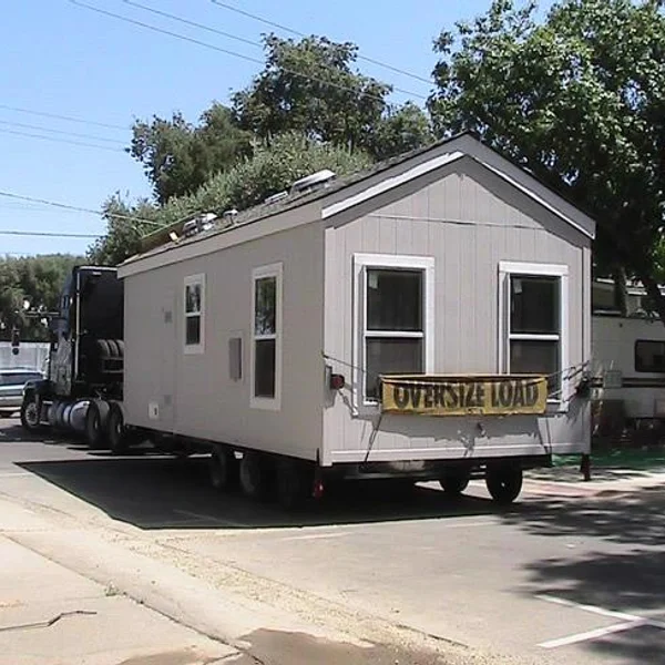 A gray ADU unit is transported as a student housing option, featuring an 'Oversize Load' sign for safe delivery.