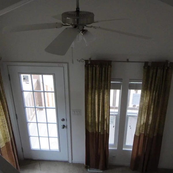 A well-lit room in this manufactured home features a ceiling fan, complementary curtains, and a glass door for outdoor access.