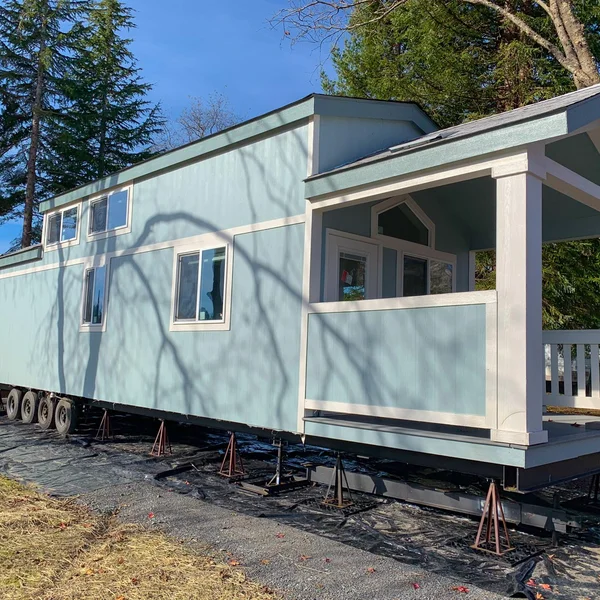 A light blue park model home sits on a trailer with leveling jacks, ready to be set up on a property.