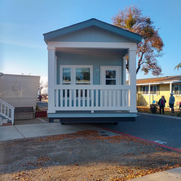 Pale blue park model tiny home with white trim and porch sits in a mobile home community.