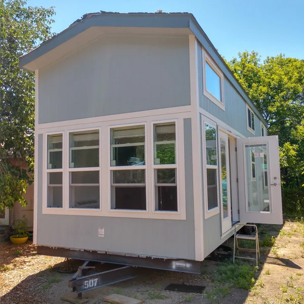 A two-story, light blue park model home with white trim sits on a trailer, showcasing a compact living solution.