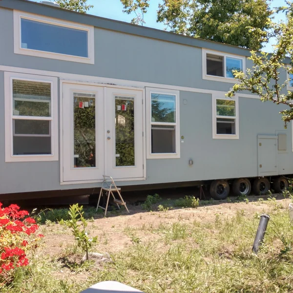 Light blue park model tiny home features multiple windows, white trim, and a double-door entrance.