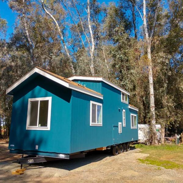 A blue park model home on wheels is set in a clearing among tall trees on a sunny day.