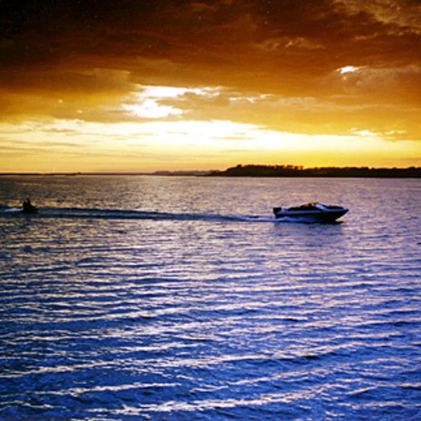 A speeding boat and jet ski cruise on the water during a vibrant sunset, near potential waterfront tiny home lot.