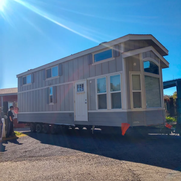 A modern gray park model home is shown on a trailer under a blue sky with visible towing equipment.