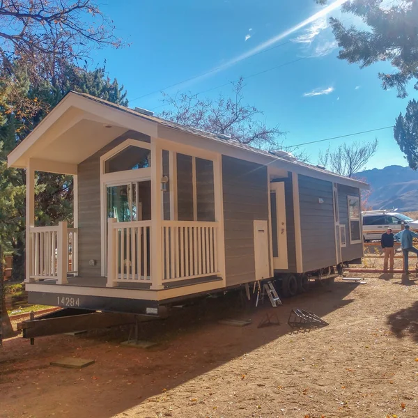 A gray park model home with a white porch is set on a trailer under a clear blue sky.