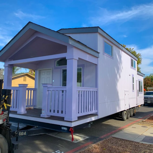 A lilac-colored park model tiny home with a covered porch is transported on a trailer under a blue sky.