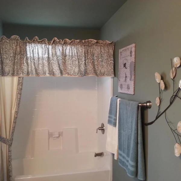 Neutral-toned bathroom in a manufactured home features a shower-tub combo, decorative art, and sage green walls.