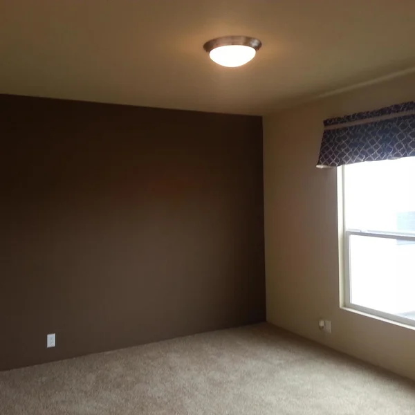 An interior of a manufactured home shows a carpeted bedroom with a brown accent wall and a window with valance.