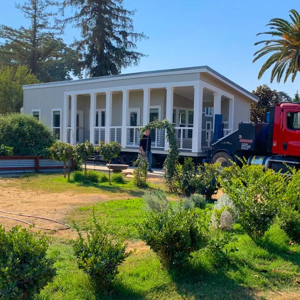 A modern, gray park model home with a porch is being delivered by a red Anderson truck.