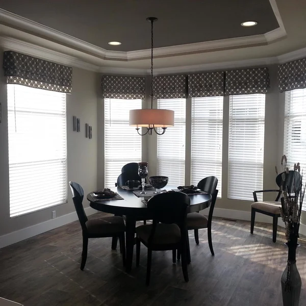 Bright dining area in a manufactured home features a round table, chairs, and large windows with patterned valances.
