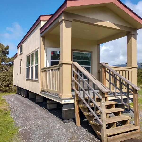 A beige park model home with a front porch and wooden steps rests on cinder blocks outdoors.