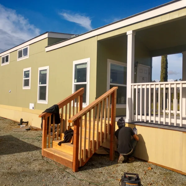 Worker installing skirting on a light green modular home with a wooden staircase and white porch railing.