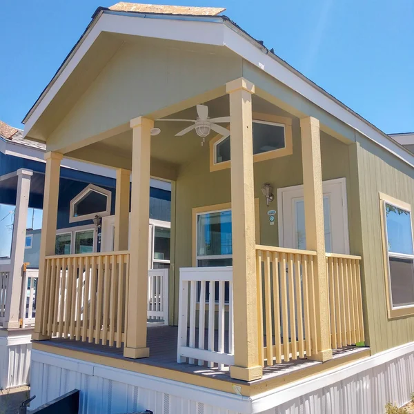 Exterior of a green park model home with a porch and yellow support columns under a bright blue sky.