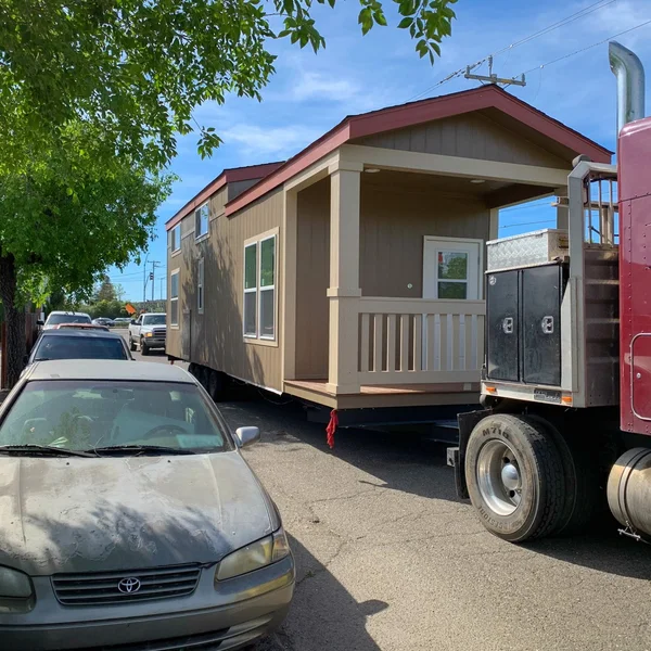 A brown park model is being transported on a trailer down a street with parked cars.