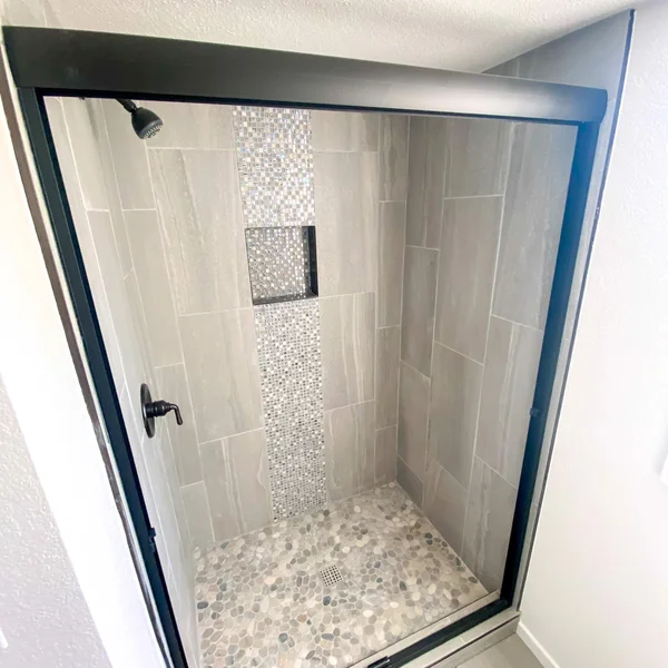 Modern ADU shower features gray stone tiles, a pebble floor, mosaic accent strip, and black trim.