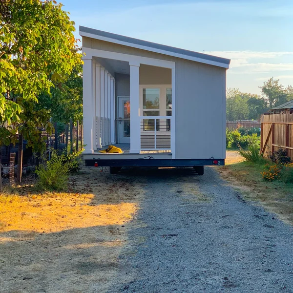 Rear view shows a modern gray park model tiny home with white porch on a gravel drive.