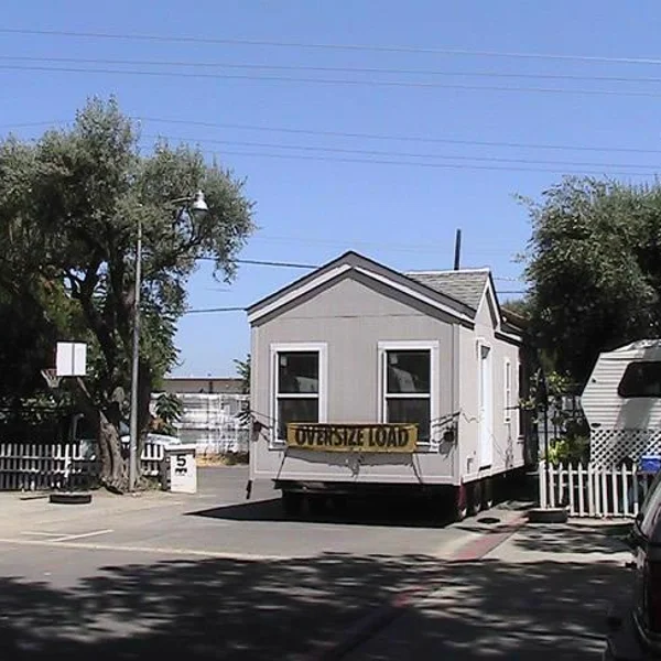 Park model tiny home, gray with white trim, is transported for student housing with an "oversize load" banner.