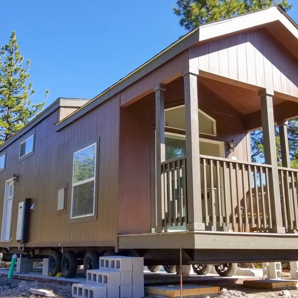 A brown park model tiny home, set on wheels with a covered porch, awaits final setup on cinder blocks.