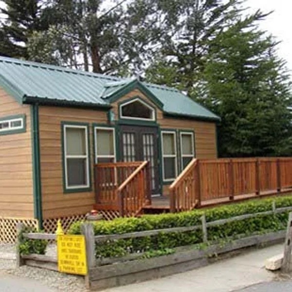 Tan park model home with a green roof features a wooden deck and is surrounded by landscaping.
