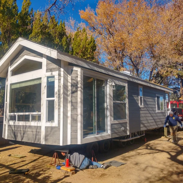 Gray park model RV sits on jacks with workers preparing the mobile home for installation.