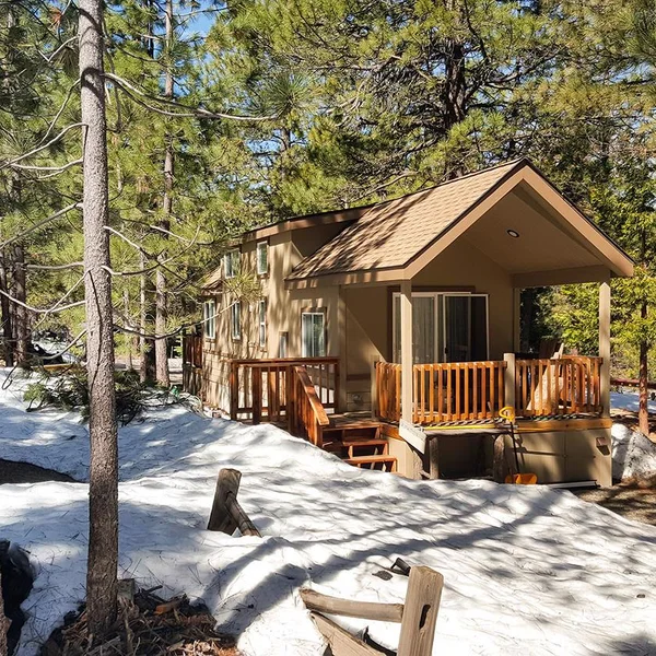 A cozy, tan park model RV nestled among pine trees in a snowy setting, featuring a wooden deck and railing.