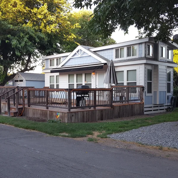 An elegant park model tiny home featuring light blue siding, a lofted design, and a large custom wooden wrap-around deck perfect for outdoor entertaining.