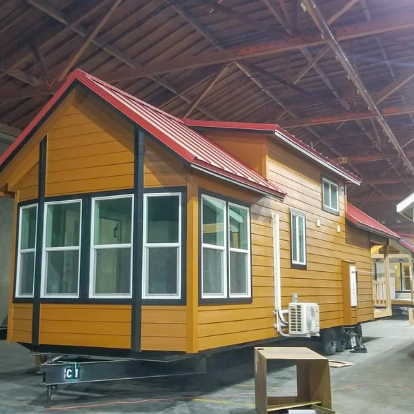 Completed tan park model tiny home with a red roof features white-framed windows, ready for delivery.