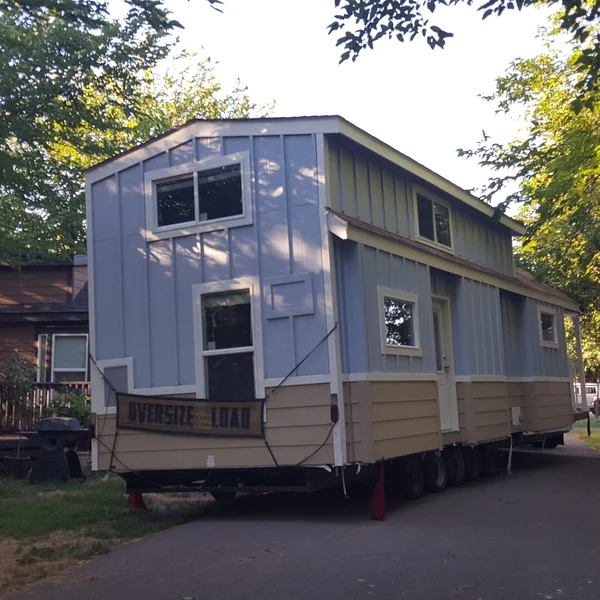 This custom-built tiny home on a trailer features a blue and beige exterior with a spacious loft design, ready for transport to its new homesite.