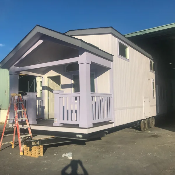 A charming white and lavender park model with a covered porch sits on a trailer, ready for transport.
