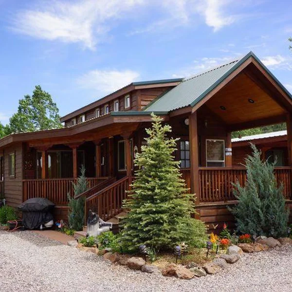 A brown, rustic park model home with a green metal roof features a wraparound porch and landscaped gravel yard.