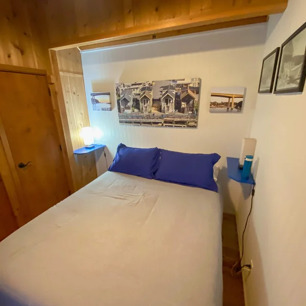 The bedroom of a tiny home features a bed with blue pillows, side tables, and coastal-themed artwork.
