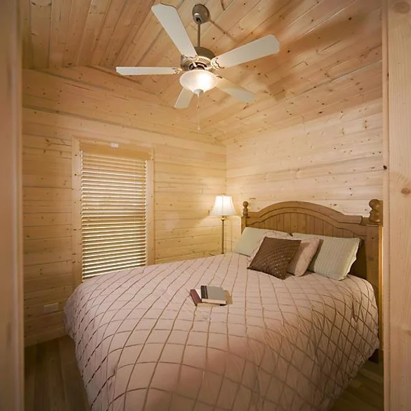 A cozy bedroom in a wood-paneled park model features a bed with a patterned quilt and a ceiling fan.