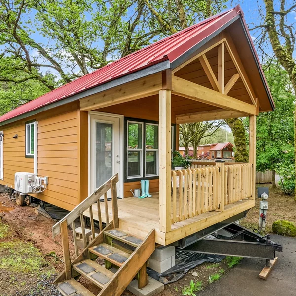 A cozy tan-sided tiny home with a red metal roof and wooden porch offers an inviting compact living space.