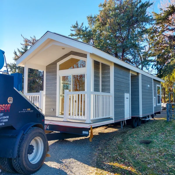 Gray park model home, with white trim and porch, is transported on a flatbed trailer.