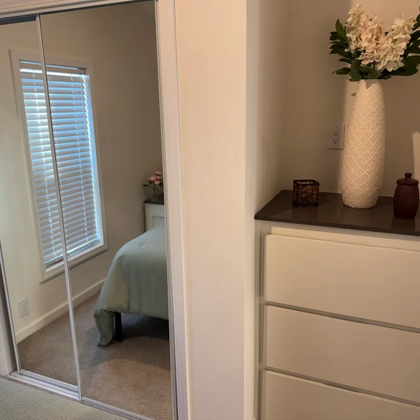 Real estate bedroom with mirrored closet reflecting window; dresser with decorative vase and floral arrangement visible.