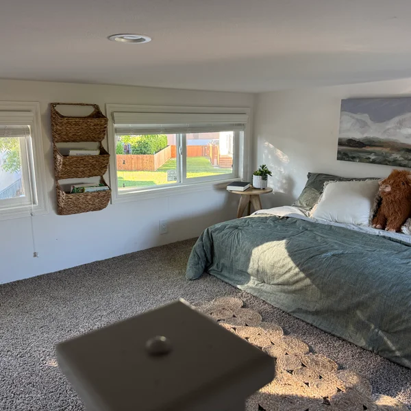Bright bedroom features a bed with sage green bedding, a white wall art, and woven wall baskets.