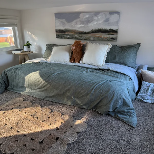 Bedroom showcasing a large bed with green comforter and white pillows, accented with a Highland cow plush and landscape art.