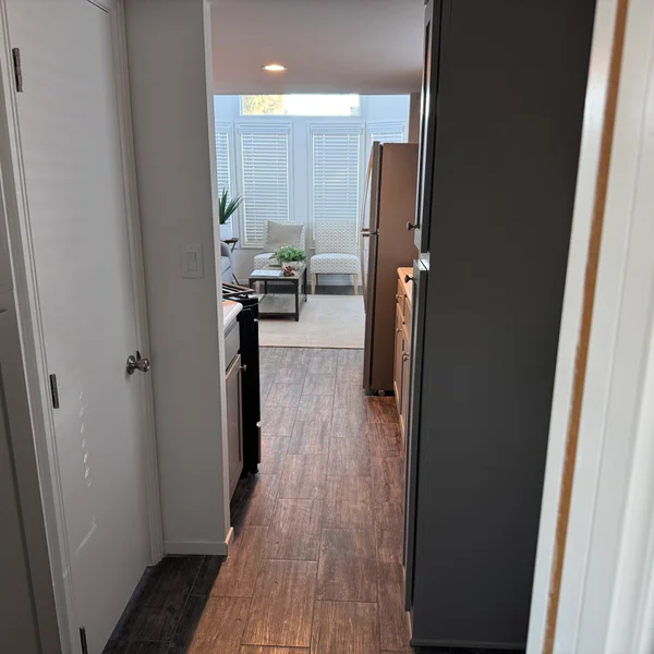 A hallway featuring wood flooring leads to a living area with white seating and a bay window.