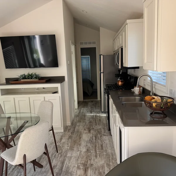 A real estate property's kitchen and dining area features white cabinets, gray countertops, and a glass dining table.