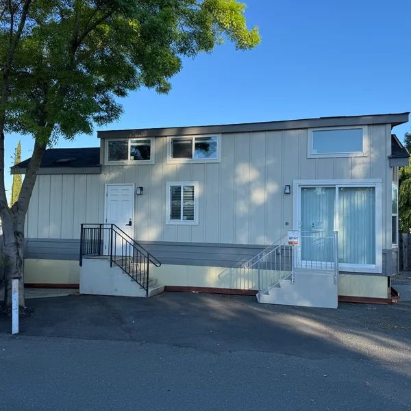 Charming tan manufactured home with grey accents, stairs, and multiple windows under a clear blue sky.