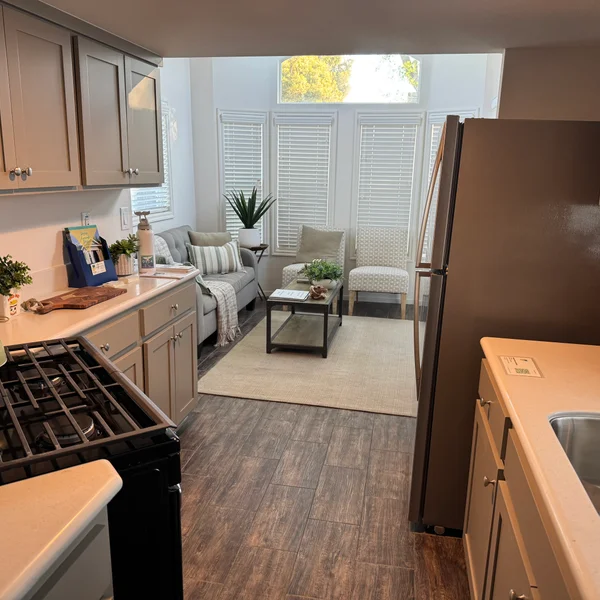 Real estate interior features a light-filled living area with gray sofa, patterned chairs, and modern kitchen.