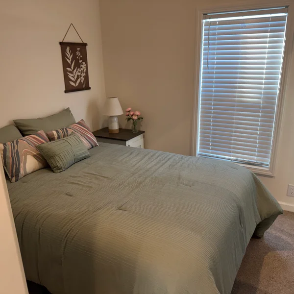 A bedroom features a green bedspread, assorted pillows, a nightstand with lamp and flowers, and a window with blinds.