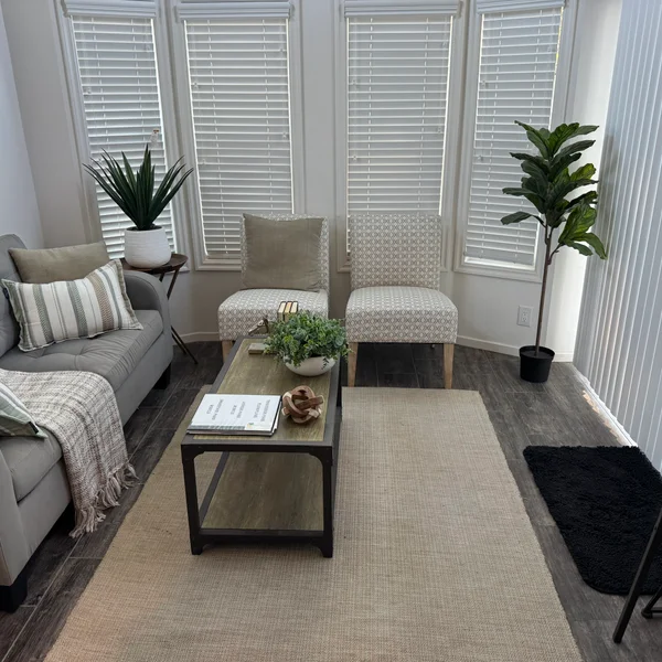 A staged living room shows a gray sofa, patterned chairs, a coffee table, and plants for a modern real estate listing.